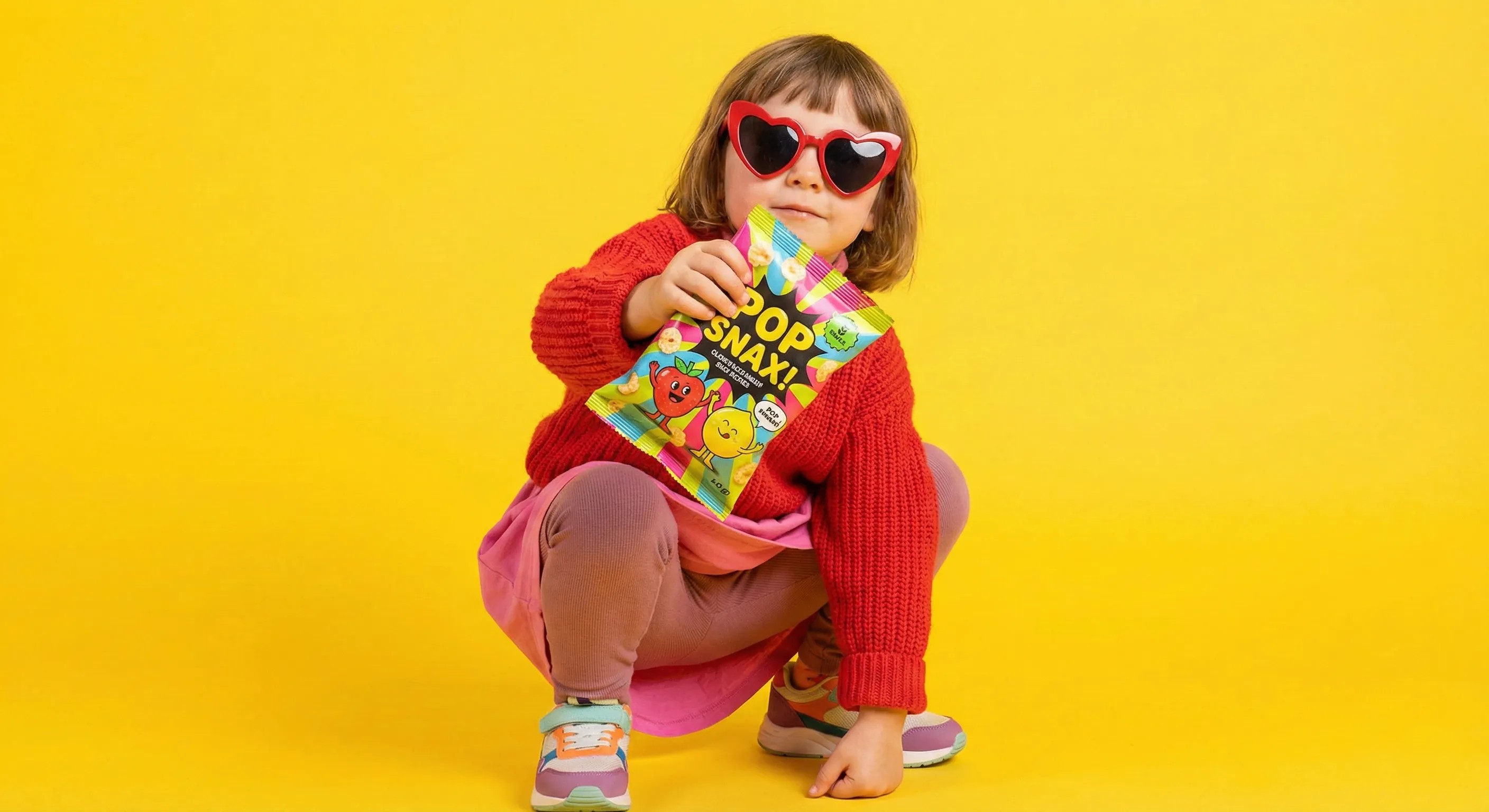 Stylish child in a red sweater and heart-shaped sunglasses squatting on a vibrant yellow background, holding a colorful snack packet toward the camera.